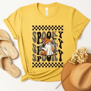 May include: A yellow t-shirt with a black and white checkered pattern and the text "Spooky S'more Spooky" in a retro font. A cartoon ghost with a pumpkin and a cup is in the center of the design.