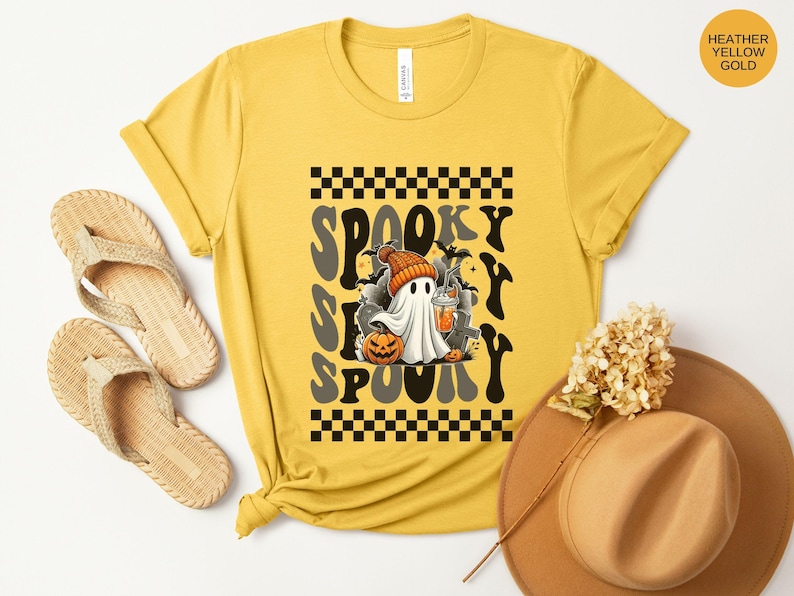 May include: A yellow t-shirt with a black and white checkered pattern and the text "Spooky S'more Spooky" in a retro font. A cartoon ghost with a pumpkin and a cup is in the center of the design.