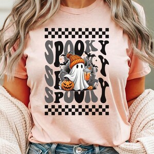 May include: A pink t-shirt with a black and white checkered pattern at the top and bottom. The shirt features a graphic of a ghost holding a beverage with the text "Spooky S  Spooky" in a retro font.