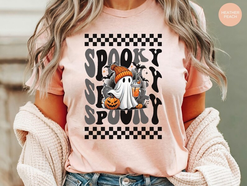 May include: A pink t-shirt with a black and white checkered pattern at the top and bottom. The shirt features a graphic of a ghost holding a beverage with the text "Spooky S  Spooky" in a retro font.