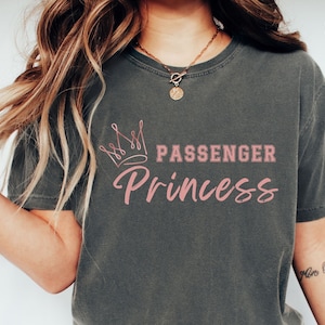 May include: Dark gray t-shirt with the text "PASSENGER Princess" in pink, topped with a crown graphic. The shirt has a vintage, washed look. The text is in a playful, feminine font.