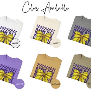 May include: A collection of t-shirts in various colors, each featuring a purple and white "Game Day Ready" graphic with a yellow bow. The colors available are white, ivory, granite, violet, mustard, and khaki.