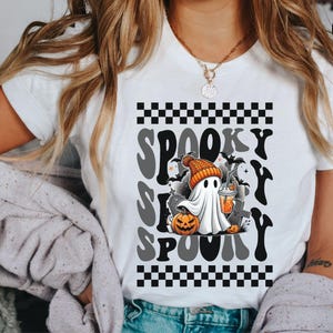 May include: White t-shirt with a black and white checkered pattern and the text "Spooky S-P-O-O-K-Y" in a retro font. A cartoon ghost wearing a hat and holding a pumpkin and a drink is in the center of the design.