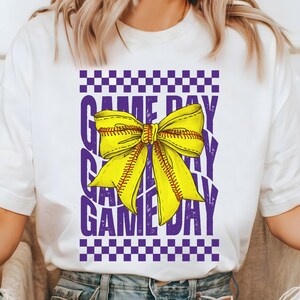 May include: White t-shirt with a purple and yellow design. The design features a softball-themed bow with the text "GAME DAY" repeated.