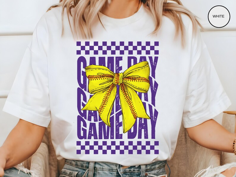 May include: White t-shirt with a purple and yellow design. The design features a softball-themed bow with the text "GAME DAY" repeated.
