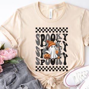 May include: A soft cream colored t-shirt with a black and white checkered pattern and a graphic of a ghost holding a pumpkin and a cup. The text "Spooky Spooky" is written in a retro font.