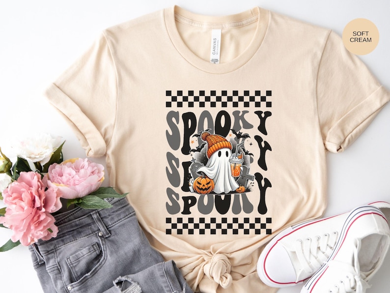 May include: A soft cream colored t-shirt with a black and white checkered pattern and a graphic of a ghost holding a pumpkin and a cup. The text "Spooky Spooky" is written in a retro font.