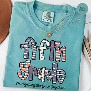 May include: Seafoam green t-shirt with the words "Fifth Grade" in a colorful design featuring fish, anchors, and crabs. The text "Navigating the Year Together" is below the main text. A silver necklace with a pendant is on the shirt.