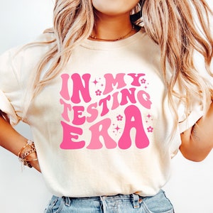 May include: A white t-shirt with a pink graphic that says "In My Testing Era" with stars and flowers around the text.