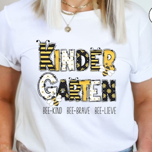 May include: White t-shirt with a black and yellow graphic that says "Kindergarten" with bees and flowers. The text below the graphic says "Bee-Kind Bee-Brave Bee-Lieve".