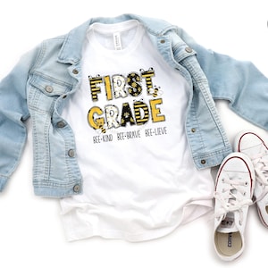 May include: A white t-shirt with the words "FIRST GRADE" in yellow and black, with bee graphics. The shirt is paired with a light blue denim jacket and white sneakers with red accents. The text "BEE-KIND BEE-BRAVE BEE-LIEVE" is below the main text.