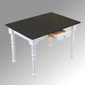 Puede incluir: Una mesa rectangular en miniatura con tapa negra y patas blancas. La mesa tiene un solo cajón que está parcialmente abierto, revelando un interior marrón claro. El cajón tiene un pequeño pomo dorado. Las patas son torneadas y tienen un diseño decorativo.
