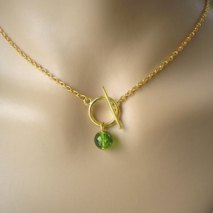 Peut inclure: Un collier doré avec un fermoir à bascule et un petit pendentif rond en perle verte. La chaîne est délicate, et le pendentif est suspendu à un anneau doré. Le collier est présenté sur un mannequin.