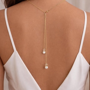 May include: A gold-coloured back necklace with a delicate chain and two dangling pendants. The pendants feature a small clear crystal and a white pearl. The necklace is worn over a white dress.