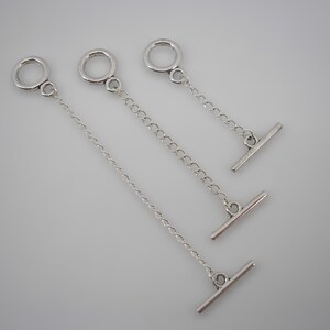 Puede incluir: Tres pinzas de corbata plateadas con diseño de barra en T. Cada pinza presenta un pequeño anillo en la parte superior, conectado a una cadena o una serie de eslabones, y termina con una barra horizontal. Las pinzas están dispuestas sobre una superficie gris claro.
