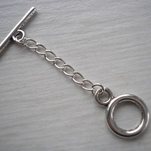 Puede incluir: Un cierre de palanca plateado, que consta de una barra en T y un anillo circular conectados por una cadena corta de eslabones ovalados. Ideal para la creación de joyas y manualidades.