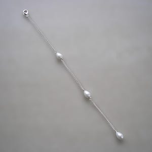 May include: A delicate silver chain bracelet featuring four teardrop-shaped pearls. The bracelet has a lobster clasp closure and is displayed against a neutral gray background. The pearls are evenly spaced along the chain.
