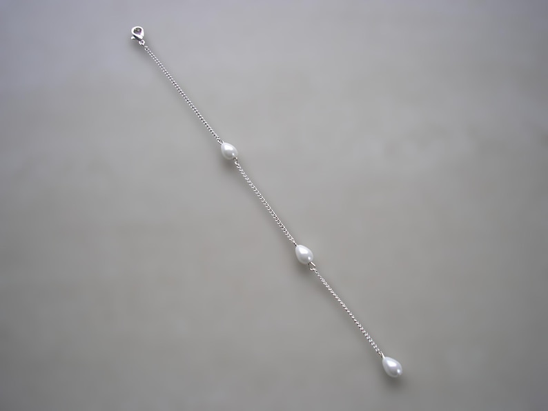 May include: A delicate silver chain bracelet featuring four teardrop-shaped pearls. The bracelet has a lobster clasp closure and is displayed against a neutral gray background. The pearls are evenly spaced along the chain.