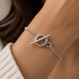 Puede incluir: Una pulsera de cadena en tono plateado con cierre de barra y argolla. La pulsera consta de una serie de pequeños eslabones metálicos uniformes conectados a un anillo circular y un componente de barra horizontal que asegura la cadena.