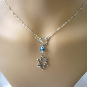 Puede incluir: Collar plateado con colgante de estrella de David. El collar tiene un cierre de palanca y una cuenta azul. El colgante es un diseño calado detallado. La cadena es delicada.