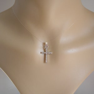 Peut inclure: Un collier avec un pendentif en forme de croix argentée orné de pierres précieuses claires et étincelantes. La croix est suspendue à un fil transparent presque invisible, créant un effet flottant. Le pendentif est présenté sur un mannequin.