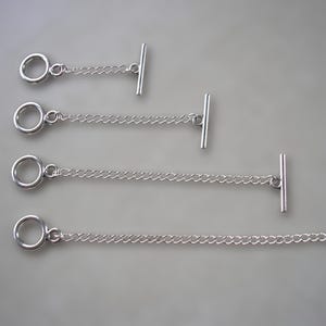May include: Four silver-toned toggle clasps arranged in a vertical row, each consisting of a circular ring, a short curb chain, and a straight bar. The lengths of the chains increase progressively from the top to the bottom of the display.