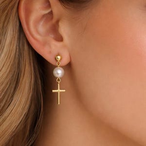 Op de afbeelding: Goudkleurige oorbellen met een parel- en kruisontwerp. De oorbellen hebben een klein gouden balletje aan de bovenkant, een witte parel in het midden en een gouden kruisbedel aan de onderkant. De oorbellen worden gedragen.