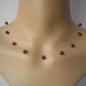 Puede incluir: Un delicado collar con pequeñas cuentas redondas de color rojo oscuro, espaciadas a lo largo de un hilo transparente casi invisible. El collar se exhibe en un maniquí, mostrando su diseño minimalista y cómo se apoya en el cuello.