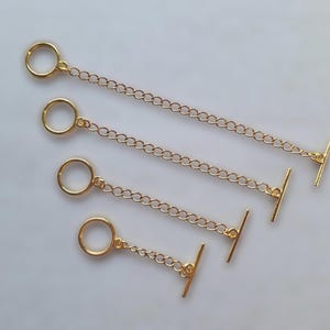May include: Four gold tie chains. Each chain features a small ring at one end and a T-bar clasp at the other. The chains are linked together. The items are displayed on a light-coloured surface.