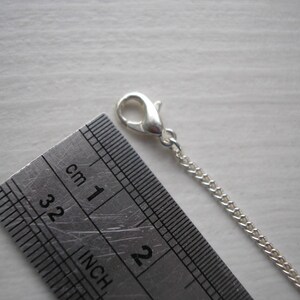May include: A close-up of a silver chain with a lobster clasp, next to a metal ruler. The ruler shows both inches and centimeters. The chain is approximately 1 inch long.