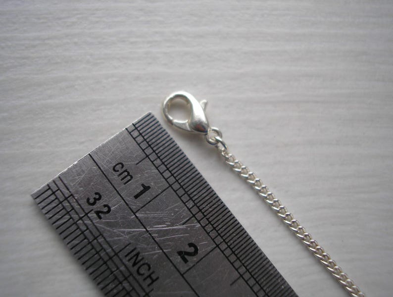 May include: A close-up of a silver chain with a lobster clasp, next to a metal ruler. The ruler shows both inches and centimeters. The chain is approximately 1 inch long.