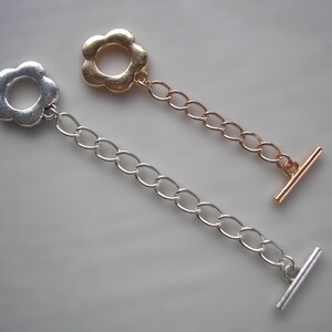 May include: Two toggle clasps for jewellery making. One is silver-coloured, and the other is gold-coloured. Each clasp features a flower-shaped loop connected to a chain and a bar. The clasps are designed to secure necklaces or bracelets.
