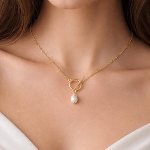 May include: A gold-toned chain necklace featuring a toggle clasp closure with a circular ring and a bar. A single white teardrop-shaped pearl pendant hangs from the bottom of the circular ring, centred at the base of the neck.