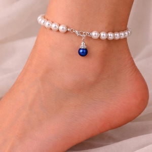 May include: Anklet with a string of white faux pearls and a silver clasp. A small, dark blue bead pendant hangs from the centre. The jewellery is worn on the ankle.