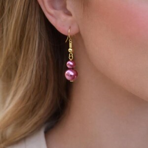 May include: Gold-tone dangle earrings featuring two pink faux pearls. The earrings have a delicate design with a small gold bead and a textured gold accent between the pearls. The earrings are shown on a person's ear.