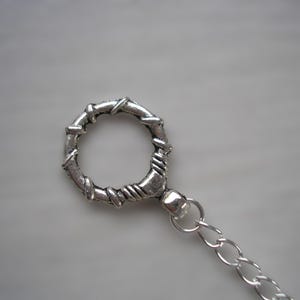 May include: Close-up of a silver-coloured metal ring with a decorative, textured design. The ring is connected to a chain. The ring has a circular shape with a detailed, woven pattern. The chain is made of small, linked rings.
