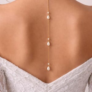 May include: A gold-toned chain necklace designed to drape down the back, featuring three white teardrop-shaped pearls spaced along the length of the chain. The necklace is worn against the skin above a white lace garment with a V-shaped neckline.