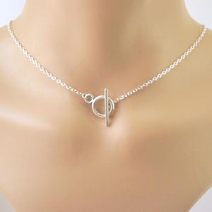 May include: A silver necklace with a toggle clasp. The necklace features a delicate chain and a circular clasp with a straight bar closure. The jewelry is displayed on a person's neck.