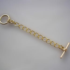 Op de afbeelding: Een goudkleurige kettingverlenger met een ring aan één uiteinde en een T-staafsluiting aan de andere kant. De ketting is gemaakt van kleine, in elkaar grijpende schakels. De verlenger is ontworpen om kettingen of armbanden langer te maken.
