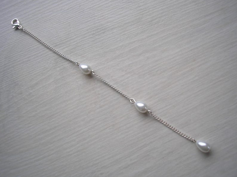 May include: A delicate silver chain bracelet featuring three teardrop-shaped pearls. The bracelet has a lobster clasp closure and is displayed on a light-colored surface. The pearls are evenly spaced along the chain.