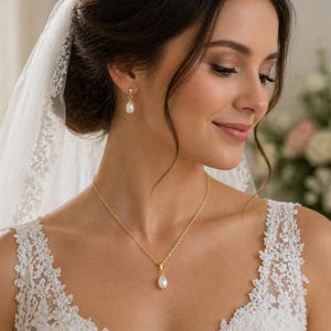 Op de afbeelding: Een bruid draagt een witte kanten trouwjurk en sluier, versierd met een gouden ketting en bijpassende pareloorbellen. De ketting heeft een fijne gouden ketting en een druppelvormige parelhanger. Zachte focus op de achtergrond met bloemen.