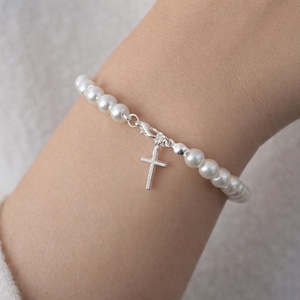 May include: A pearl bracelet with a silver cross charm. The bracelet features a strand of white, round pearls and a small, silver cross pendant. The clasp is also silver. The bracelet is worn on a wrist.