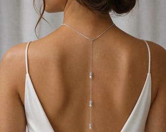 May include: A silver-toned back necklace featuring a delicate chain that drapes down the spine. The design includes three evenly spaced cylindrical metal accents, each paired with a small, round white pearl bead, ending in a single pearl drop.
