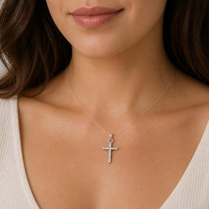 Pode incluir: Um colar com pingente de cruz prateada com pedras transparentes e brilhantes. A cruz é suspensa por uma corrente delicada, criando um efeito flutuante. O colar é usado sobre uma blusa creme.