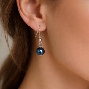 May include: A single dangle earring with a dark blue pearl. The earring has a silver-colored hook and chain. The pearl is a deep, lustrous color, and the earring is shown against a blurred background.