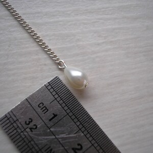May include: A silver chain necklace with a pear-shaped, off-white pearl pendant. A ruler is placed next to the pendant, showing measurements in centimeters. The pearl is approximately 1 cm in length.