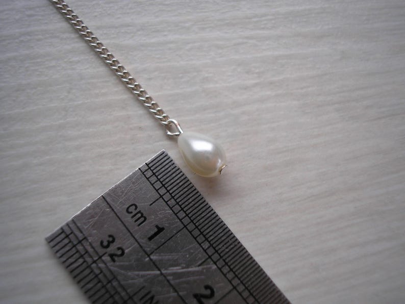 May include: A silver chain necklace with a pear-shaped, off-white pearl pendant. A ruler is placed next to the pendant, showing measurements in centimeters. The pearl is approximately 1 cm in length.