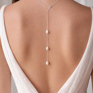 Peut inclure: Un collier en argent délicat avec un design en forme de Y, orné de trois perles blanches pendantes. Le collier est porté dans le dos, mettant en valeur le décolleté d'une robe blanche. Un bijou simple et élégant.