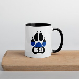 Könnte beinhalten: Weiße Keramiktasse mit schwarzem Henkel und Rand. Die Tasse zeigt ein blaues und schwarzes Pfotenabdruck-Design mit dem Text "K9" in Weiß. Die Tasse steht auf einer Holzoberfläche.