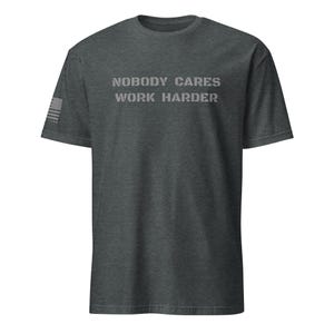 Puede incluir: Una camiseta gris oscuro jaspeada con el texto "Nobody Cares Work Harder" impreso en blanco en la parte delantera. Una pequeña bandera estadounidense es visible en la manga izquierda.